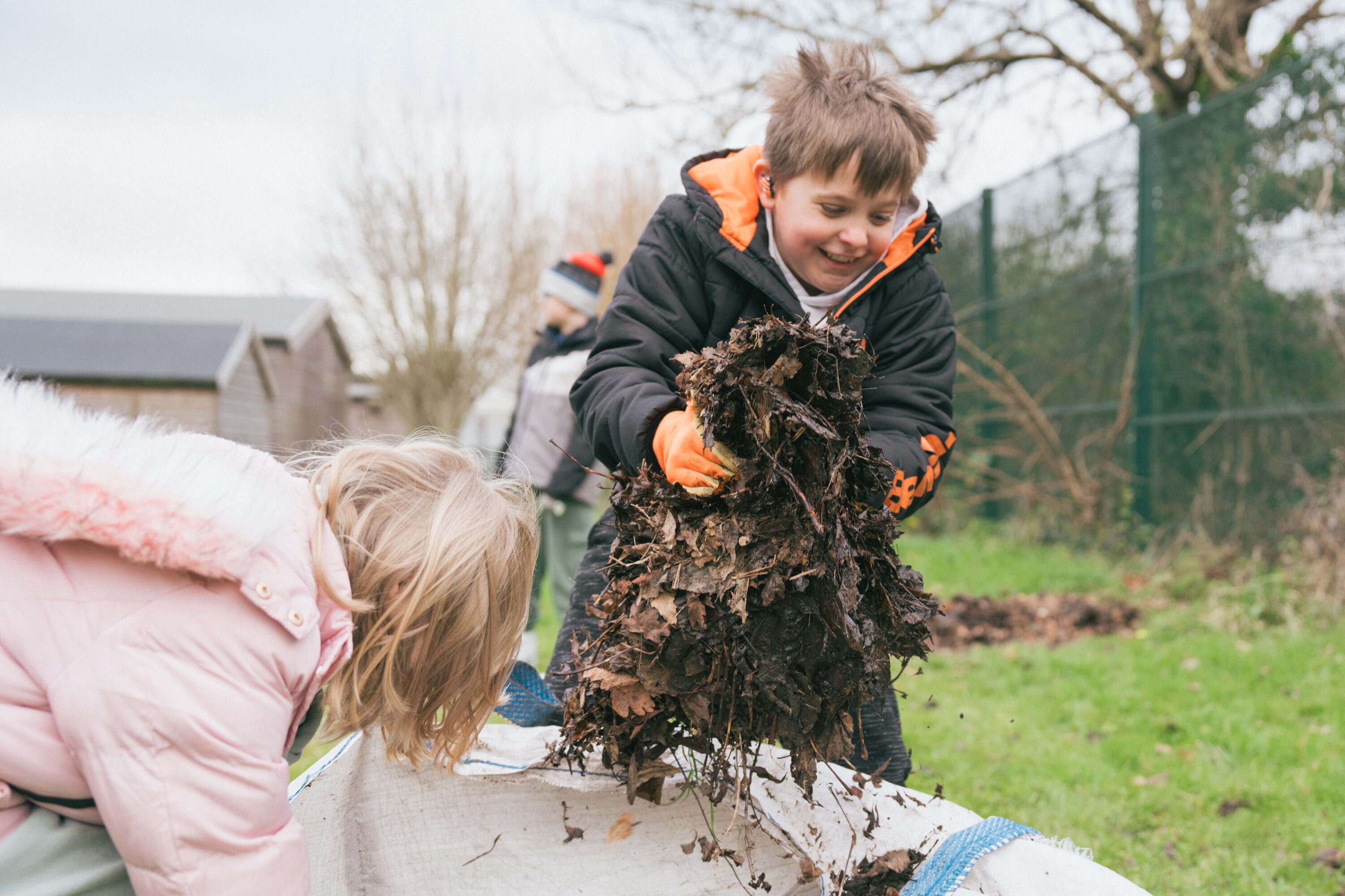 A child smiles as they grab a big handful of leaf mulch from a white bag. They are outdoors.