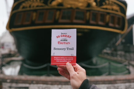 A leaflet of a sensory trail is being held up against the background of SS Great Britain boat