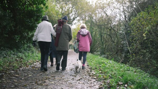 A group of people walk along an informal outdoor path away from the camera there is also a white dog