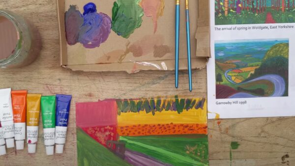 Colourful paint brushes, paints and paintings laid out on a table