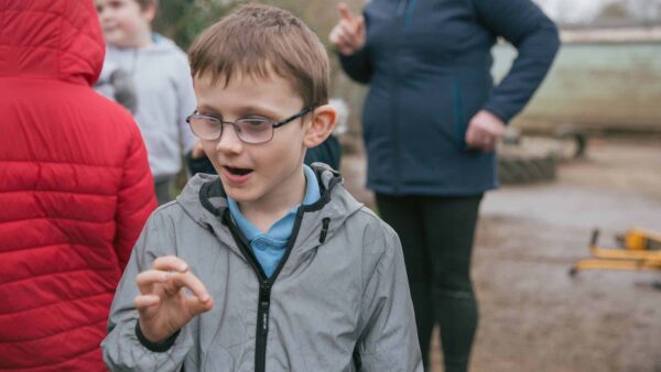 A child with glasses and a grey coat signs in Makaton. They are outside with adults behind