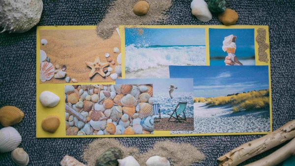 A handmade postcard with images of the beach and sea. The postcard is surrounded by sand, shells and driftwood