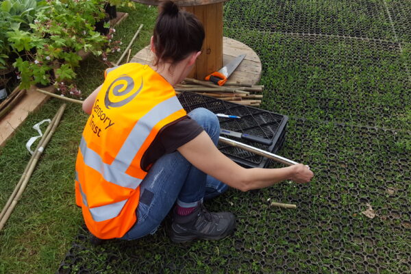 Person wearing an orange Sensory Trust branded vest has back to camera outdoors measuring