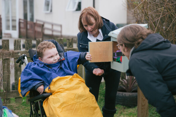 A child in a wheelchair opens the door of a bird box with two adults in a garden