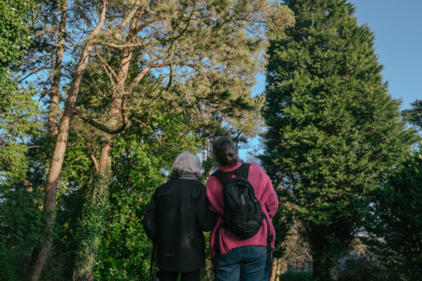 Two people stand side by side looking up at some tall trees