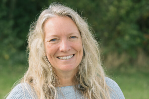 Laura looks at the camera and smiles. She is outdoors with greenery behind her and out of focus