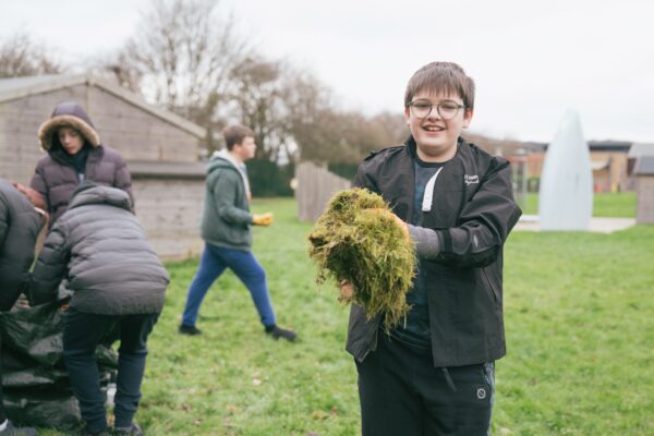 A child is outside and holding a handful of grass clippings they smile at the camera. Behind them other people are busying around