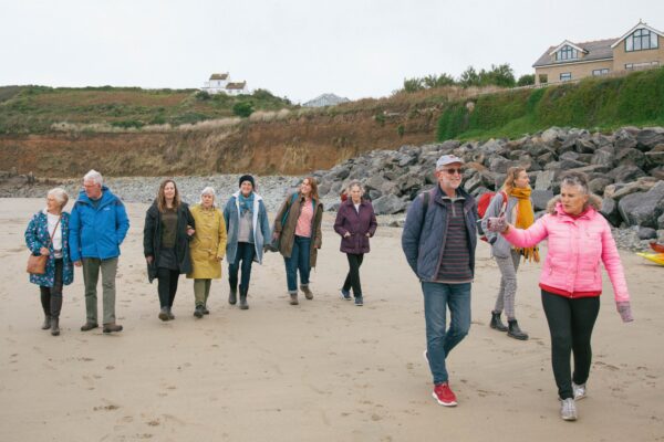 A group of people of various ages are walking along a beach they are pointing things out and smiling