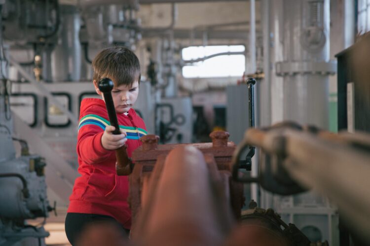 Child holds a long metal pole whilst exploring some machinery indoors