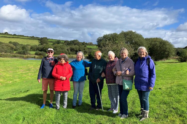 A group of people gather outdoors by a lake. It is cold but the sun is shining