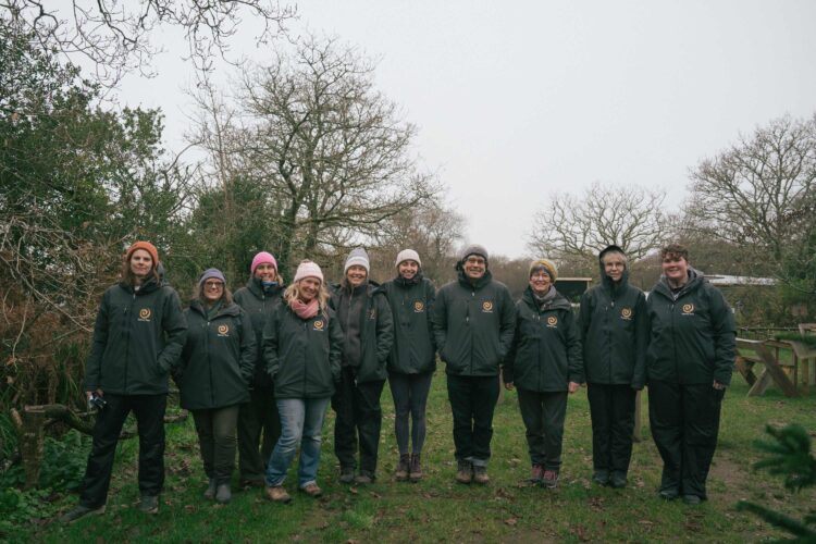 A group of ten people are standing outdoors all dressed in matching black Sensory Trust branded coats. They are wearing woolly hats, it is a grey day