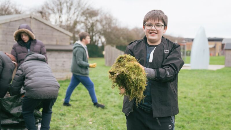 A child is outside and holding a handful of grass clippings they smile at the camera. Behind them other people are busying around
