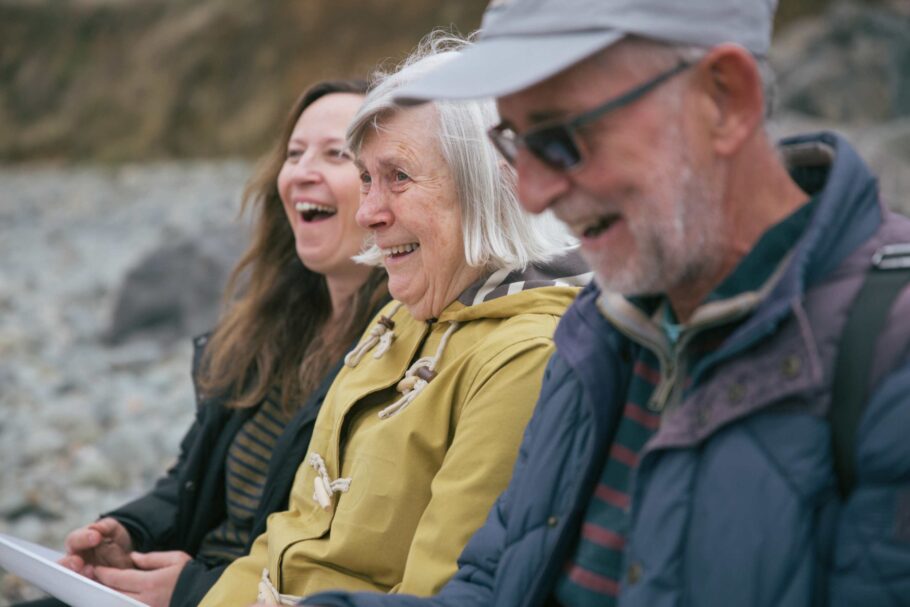 Three people sit on a stony beach in Autumn they are laughing amongst themselves